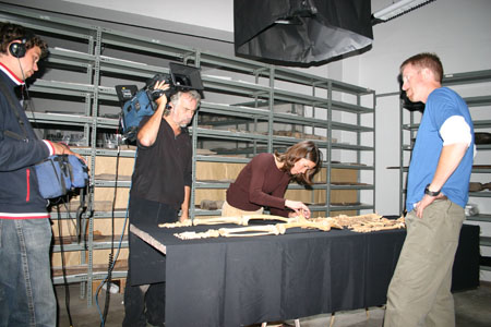 Tiffiny is hunched over a skeleton. Ken, Peter, and AJ are standing around the table. Ken is wearing a blue t-shirt, long sleeved white shirt, and khaki pants. AJ (on the left) is wearing jeans and a blue jacket with white and red on the bottom, collar, and sleeves. He is holding a camera in a blue bag. Peter is wearing a black sweater and dark jeans. His camera is over his shoulder.