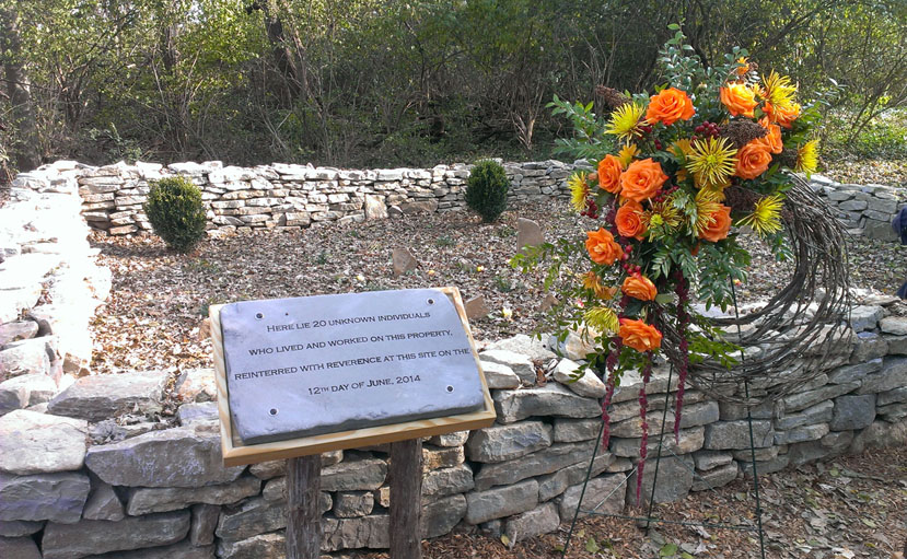 There is a small rectangular stone wall with headstones inside the barrier. There is a plaque that says "Here lies 20 unknown individuals who lived and worked on this property, reinterred with reverence at this site on the 12th day of June, 2014" next to a wreath with orange and yellow flowers.