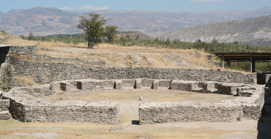 A short wall or fence-like structure made of stone is in a D shape in an open grassy area.There are mountains in the background.