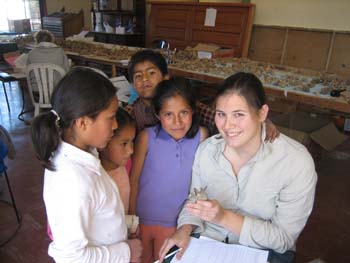 Ellen with four kids around her. She is showing them a vert. Two of the kids are looking at it and the other two are looking into the camera. Ellen is looking into the camera. She has paper in her lap and is looking into the camera. There is a table covered with verts behind her and another with other bones. Ellen is wearing a long sleeved white shirt and has brown hair pulled back.