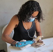 Kristina is wearing a black tanktop, glasses, a blue mask, and white gloves. She has curly black hair. She is holding a drill in one hand and a jaw bone in the other. She is looking down at them.She is sitting in a white chair and a white table.