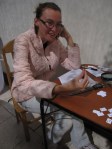 Alysha is holding up a tooth and smiling into the camera. There is a printed out spreadsheet and a gray ruler on the table in front of her on a wooden table. There is a pile of small white papers and some are laying separately with writing on them. The wall is white and so are the tiles on the floor. Alysha is wearing a pink jacket, gray pants, and glasses. Her brown hair is pulled back.