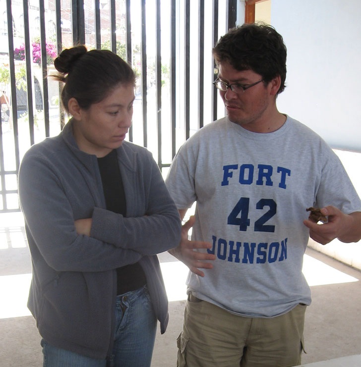 Mirza and Roberto are talking. They are standing in the shade but the sun is coming through a black metal gate behind them. Mirza is wearing a black tshirt, gray jacket, and jeans. Her brown hair is in a bun. Roberto is wearing a gray shirt that says in blue lettering, "Fort; 42; Johnson" as well as kahki pants. He wears glasses. He has short brown hair.