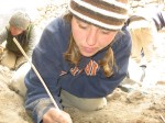 A close of up Sara crouching by an excavation that can't be seen. Cat is in the back on the left and someone's back is on the right. Sara is holding a small wooden tool into the hole. She is wearing a blue sweatshirt with orange letters with white outlines. She is also wearing khaki pants and a brown, black, and white beanie. She has a piercing in the eyebrow on the left. Her brown hair is in pigtails. In the background, Cat is wearing a gray shirt, a green vest, khaki pants, and a gray beanie. The other person is wearing a blue swearshirt and khakis.