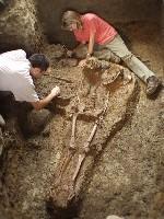 Carrie Anne is sitting down next to a skeleton that is partially excavated from the dirt. She is looking down and to the right. There is another person wearing a white shirt next to the skeleton. Carrie Anne is wearing a pink tshirt, khaki pants, and brown shoes. Her blonde hair is shoulder length.