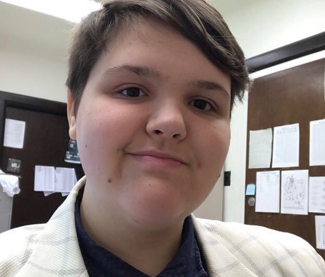 Terrance is smiling into the camera. He is wearing a blue collared shirt and a white suit jacket with gray lines making a square pattern. He has short brown hair. There are two white walls making a corner. Both of the walls have wooden doors with printouts on them.