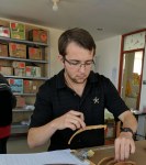 Tom is looking at a rib he is holding. There are more ribs and a clipbaord on the wooden table in front of him. He is wearing a black polo with the Vanderbilt star on it- black star with a golden outline and a white V- a black watch, glasses, and has one black ear bud in his ear. He has short brown hair as well as brown facial hair.