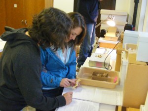 Teresa and Kelly are looking at a bone that Teresa (left) is holding. There are papers and plastic trays with bones on the white counter in front of them. Teresa is wearing a blue jacket and dark pants. She has shoulder length brown hair. Kelly is wearing a black hoodie and has chin length black hair.