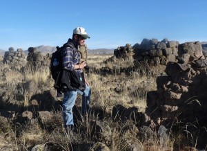 Matt is standing in the grass around several circular structures made of stone. He is facing left. He is wearing a blue and white plaid shirt, jeans, glasses, a white baseball cap with a green bill, glasses, a black back pack on his shoulders, and another black back over the shoulder closest to the camera.
